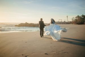 A bride and groom walk on a beach at sunset